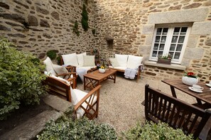 A stone-walled courtyard with wooden furniture, including sofas and a table, and a window with a flower box. - Demeures & Châteaux - Hôtel de la porte Saint-Malo - DINAN (Dinan)