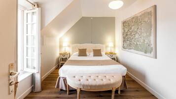 A bedroom with a large bed, two bedside tables with lamps, a bench, a window, and a painting on the wall.