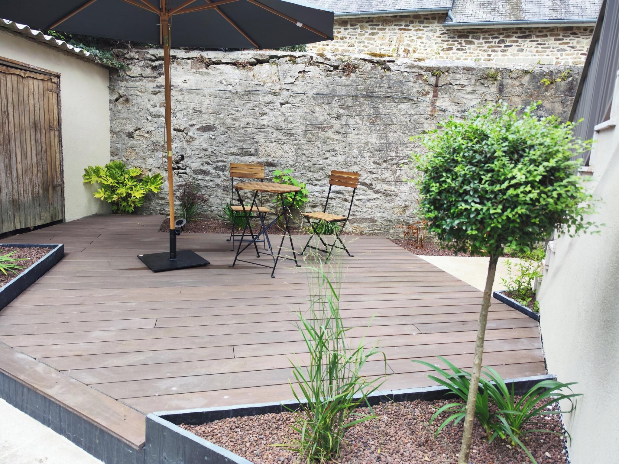 Une terrasse avec un sol en bois, deux chaises en bois, une table et un parasol noir.