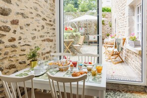 A breakfast table set with plates, cups, and a basket of bread, with a stone wall and a view of the garden outside. - Demeures & Châteaux - Hôtel de la porte Saint-Malo - DINAN (Dinan)