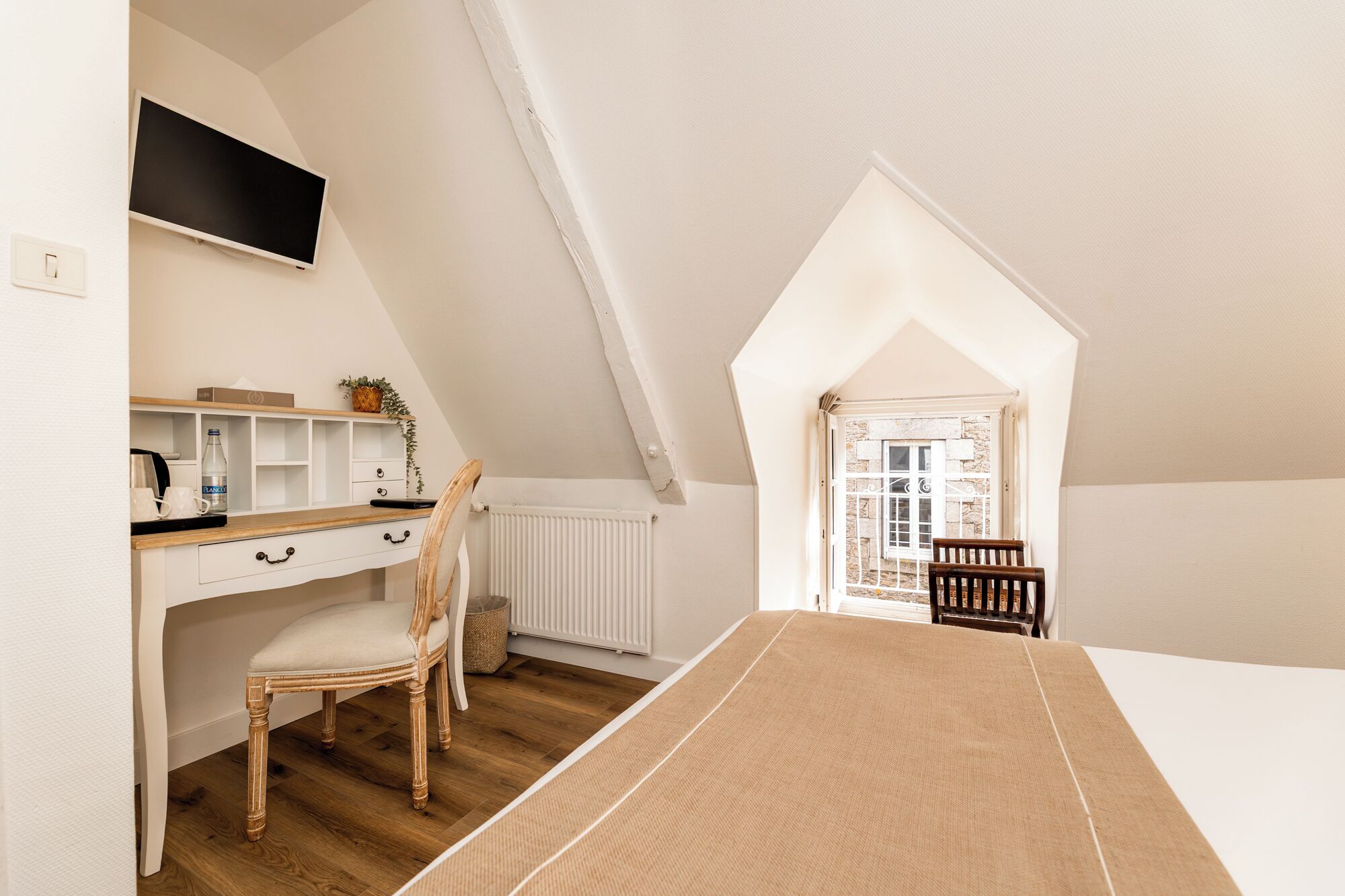 Ein Schlafzimmer mit schrägem Dach, einem Holztisch, einem Stuhl, einem Bett und einem Fenster mit Blick nach draußen.