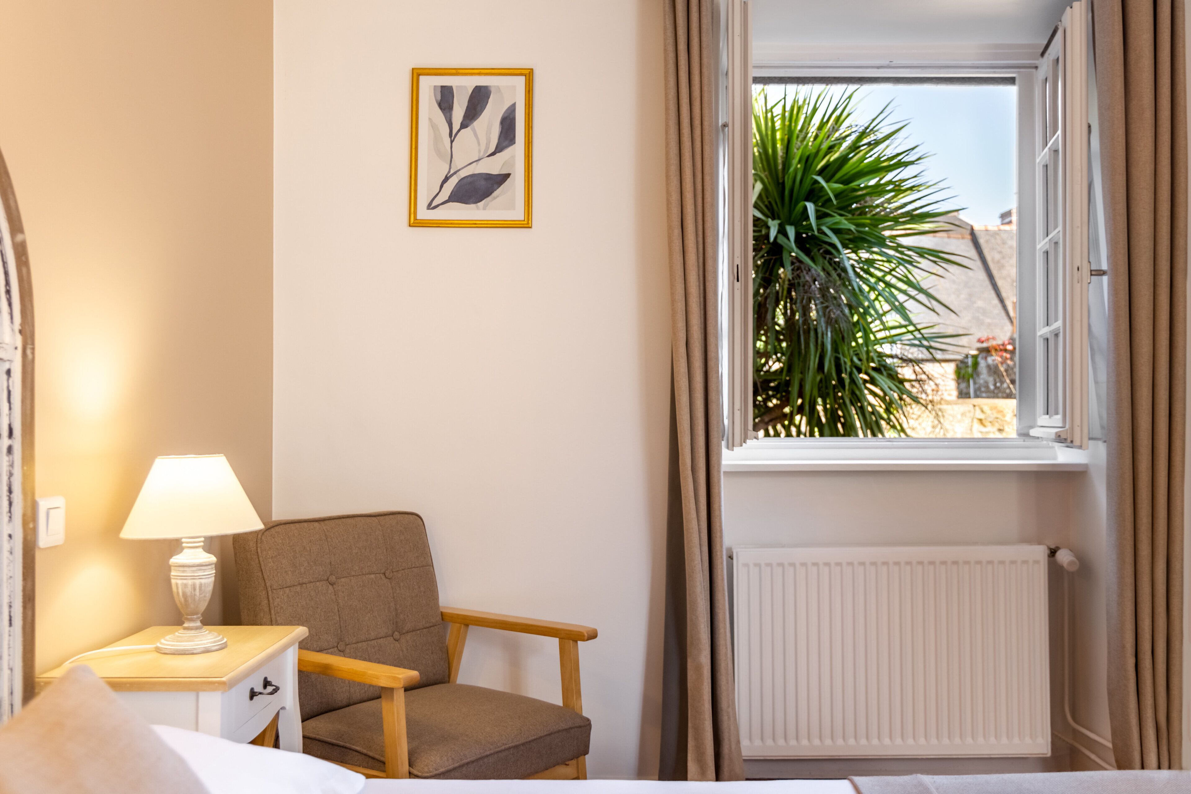 A hotel room with a bed, a bedside table with a lamp, a chair, a window with curtains, and a radiator.
