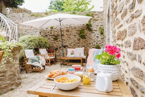 A stone-walled outdoor patio with a wooden table set for breakfast, including a bowl of cereal, a glass of juice, and a white pitcher. - Demeures & Châteaux - Hôtel de la porte Saint-Malo - DINAN (Dinan)
