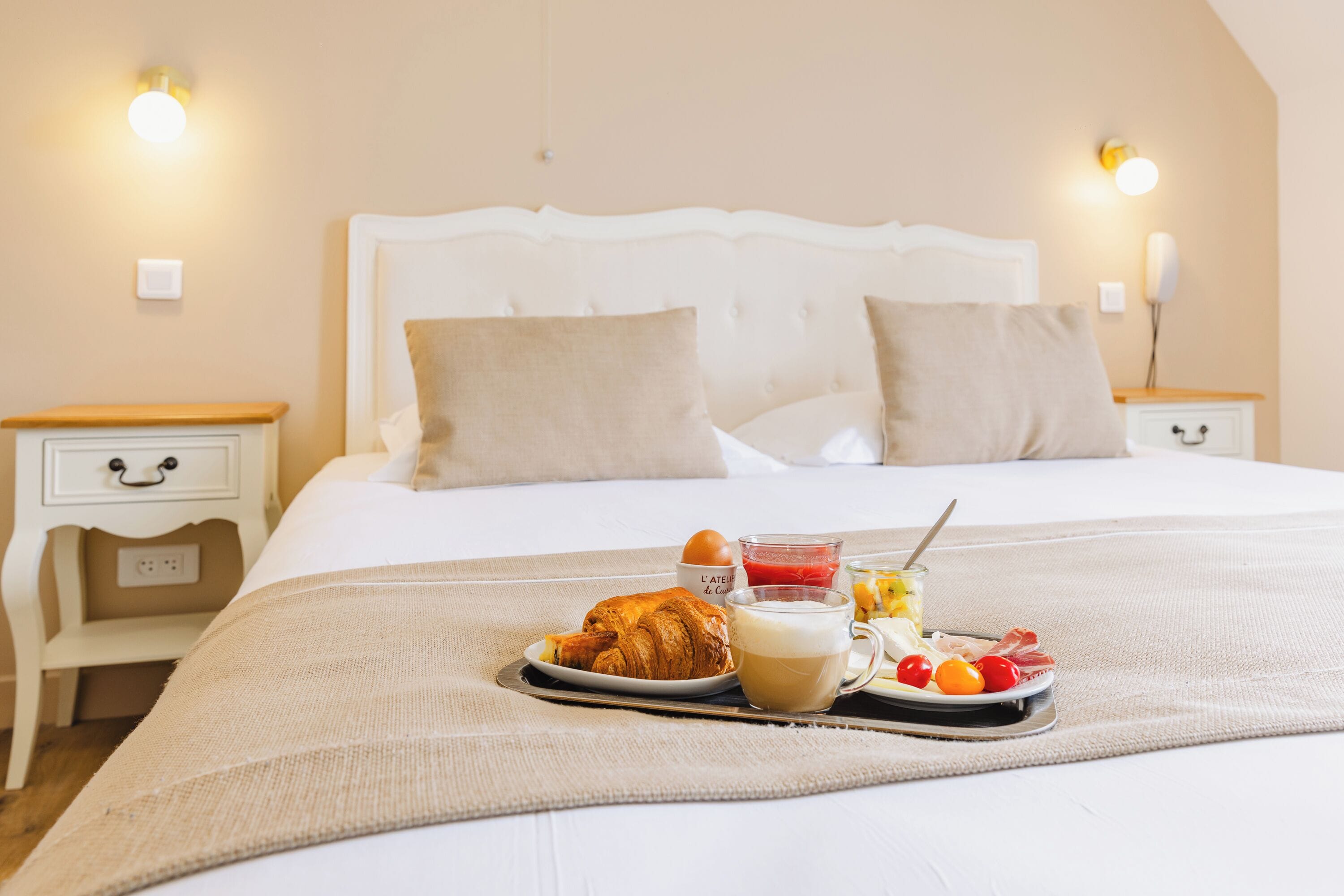 Una cama impecable con una bandeja de desayuno, que incluye un croissant, café y fruta, en una habitación de hotel con un diseño sencillo y limpio y dos lámparas de noche.