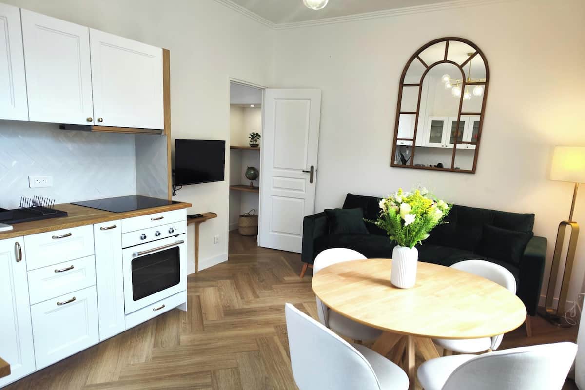 A modern kitchen with white cabinets, a wooden countertop, and a built-in oven. There is a dining area with a round table and four chairs. A black sofa and a mirror are visible in the background.