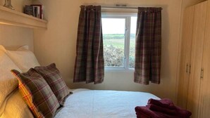 A bedroom with a bed, pillows, a window with curtains, a wardrobe, and a shelf with books. - Cosy, Quiet Static Caravan, Mawgan Porth, St Eval (Wadebridge)