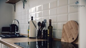 Cozinha com bancada preta, azulejos brancos, tábua de corte de madeira, uma garrafa e utensílios de cozinha.