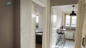 Corredor que dá para uma área de refeições com mesa e cadeiras, janela com vista e uma cama ao fundo.