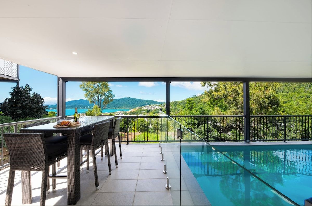 A balcony with a pool, dining table, and chairs, overlooking a mountainous landscape.