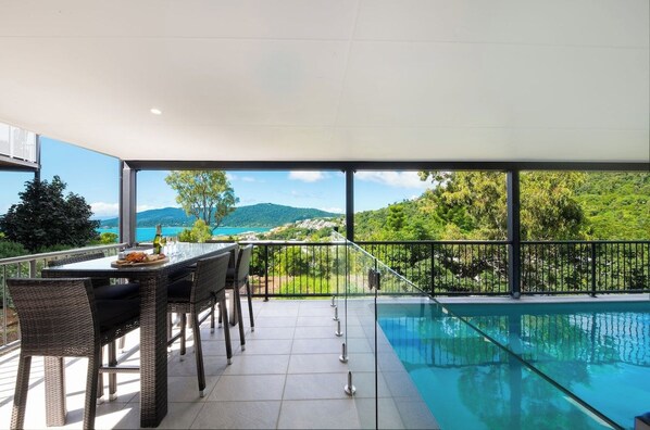 A balcony with a pool, dining table, and chairs, overlooking a mountainous landscape.