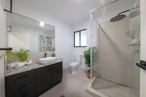 A modern bathroom with a glass-enclosed shower, a white sink, dark cabinets, and a large mirror.