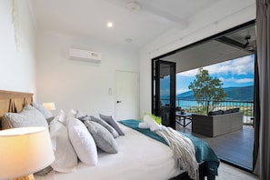 A bedroom with a bed, pillows, a bedside lamp, and a view of a balcony with a table and chairs.