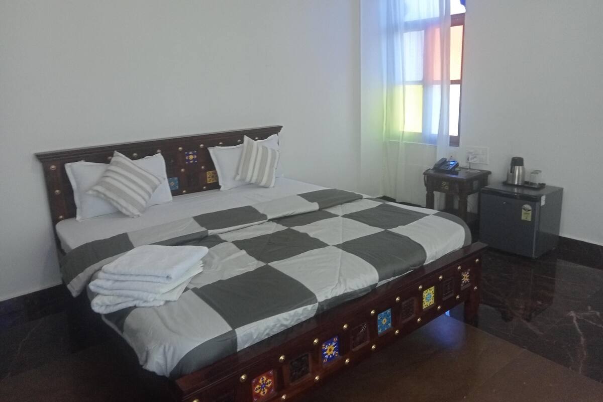 Un lit bien fait, recouvert d’une couvre-lit à carreaux, une table de chevet avec une lampe et un petit réfrigérateur.