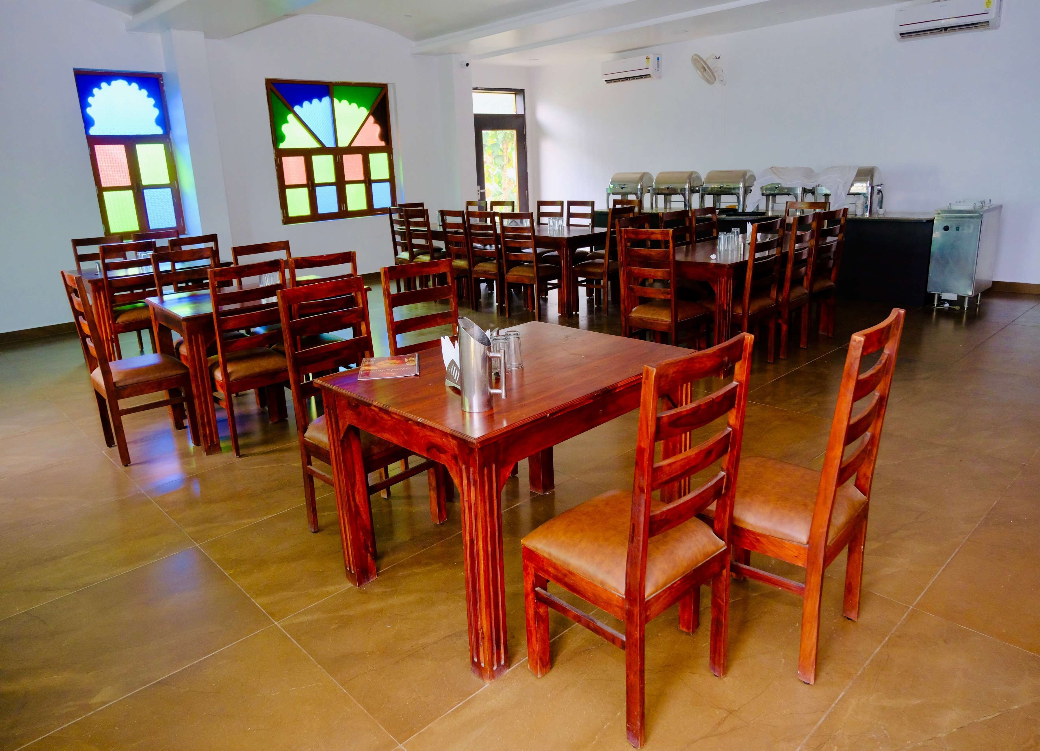 Un comedor amplio con varias mesas y sillas de madera, unidades de aire acondicionado y un ventanal grande con vidrieras de colores.