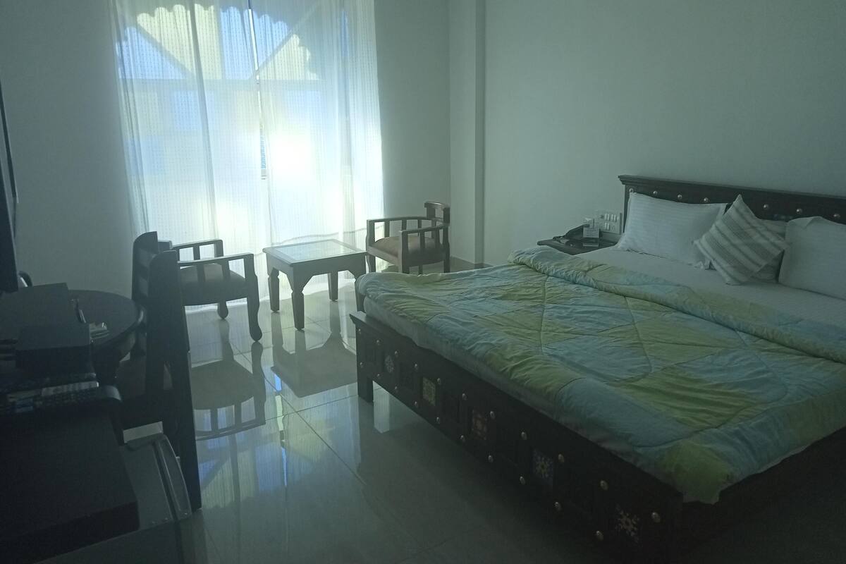 Une chambre d’hôtel avec un lit, une petite table, deux chaises, une table de chevet avec une lampe et une fenêtre avec des rideaux légers.
