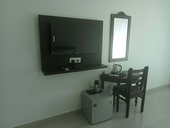 A small room with a TV mounted on the wall, a small table with a kettle, a chair, and a mini-fridge.