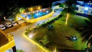 Vista aérea de un hotel con piscina, zona de estacionamiento y jardín bien iluminado con asientos.