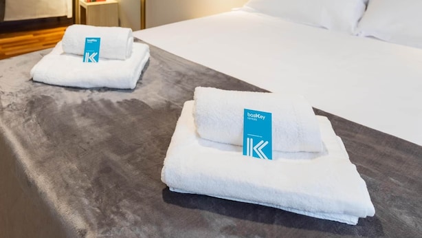 Habitación de hotel con cama, dos toallas con un logotipo, una mesita de noche, una lámpara de pared y un cuadro enmarco.