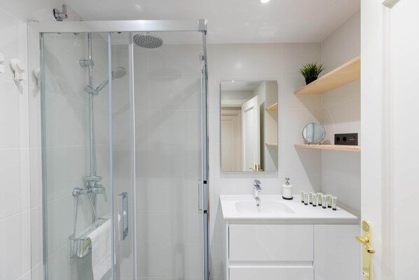 Un baño moderno con ducha envuelta en vidrio, un lavamanos blanco con artículos de tocador y un espejo sobre el lavamanos.