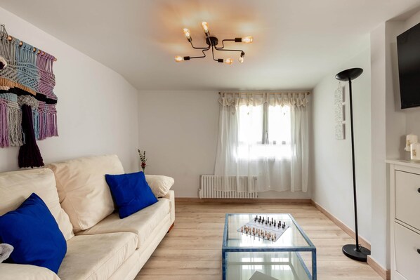 A living room with a beige sofa, blue cushions, a glass coffee table, a macrame wall hanging, a modern chandelier, a floor lamp, and a TV on the wall.