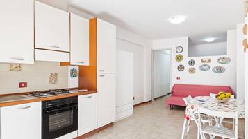Cozinha com armários brancos, fogão a gás e forno embutido. Há uma área de jantar com mesa e cadeiras, e um sofá vermelho.