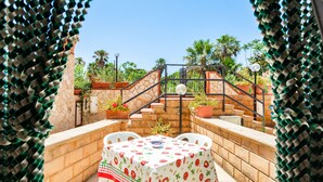 An outdoor patio with a table covered in a tablecloth with a fruit pattern, surrounded by stone walls and potted plants.
