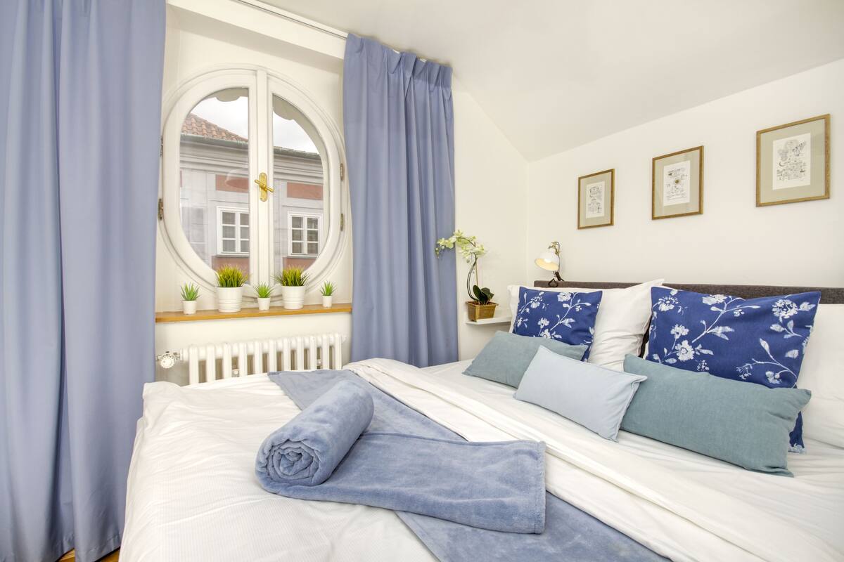 A neatly made bed with a blue towel, a window with curtains, and three framed pictures on the wall.
