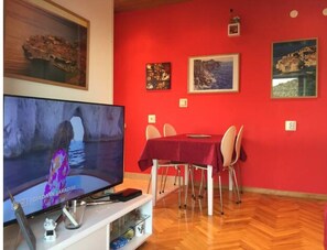 A room with a red wall, a flat-screen TV, a dining table with chairs, and artwork.