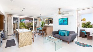 A modern living room with a glass coffee table, a grey sofa with blue cushions, and a dining area with a glass table and chairs. - Casa Turquesa - Private Pool! Local Neighbourhood (Puerto Vallarta)