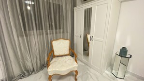 Quarto com guarda-roupa branco, cadeira de madeira, mesa de vidro e cortina cinza.