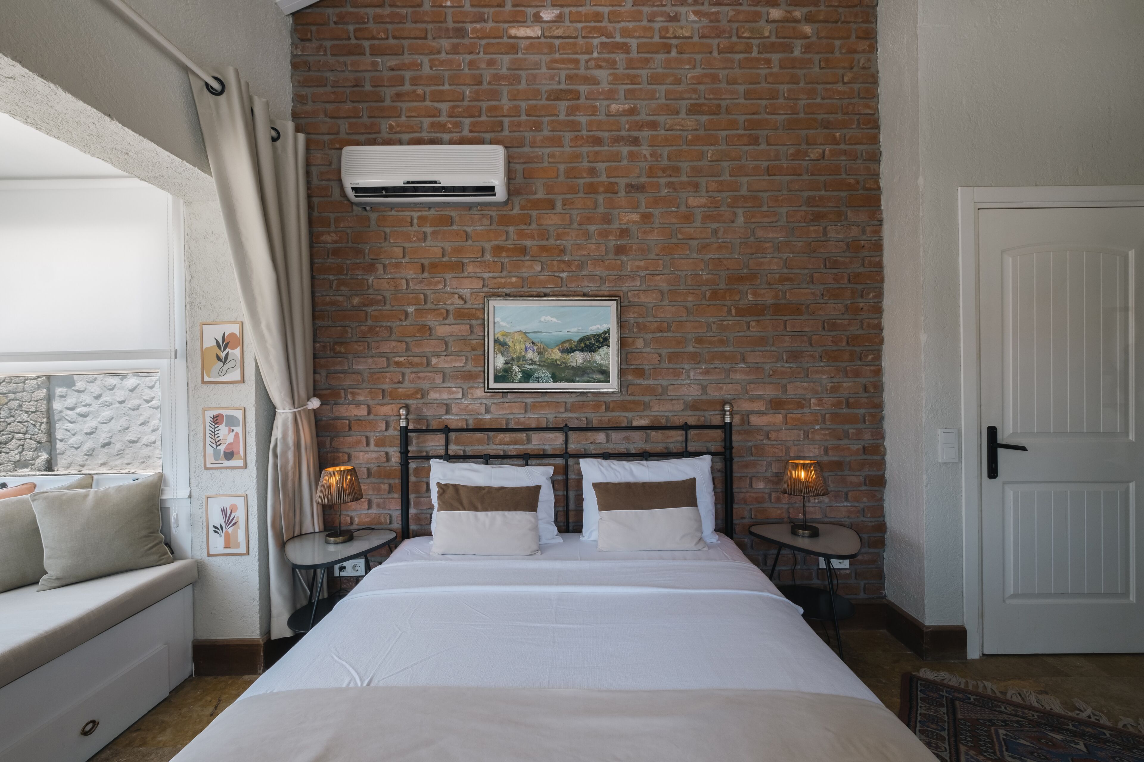 A bedroom with a brick wall, a bed with white bedding, a nightstand with a lamp, and a framed picture on the wall.