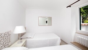 Une petite chambre d’hôtel blanche, composée d’une seule pièce, avec un lit, une table de chevet, une lampe et une fenêtre donnant sur un paysage verdoyant.