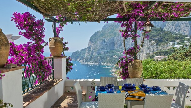 Une terrasse meublée de mobilier en osier blanc, une table dressée avec de l’assiette bleue et jaune, et de grands vases ornés de fleurs violettes.