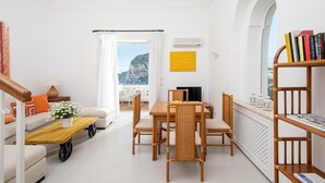 Un salon lumineux et moderne, doté d’un canapé, d’une table basse et d’un coin repas. Un balcon avec vue sur la mer et les montagnes est visible à travers de grandes fenêtres.