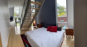 Una camera da letto moderna con un letto grande, una scala in legno e vista su un ponte e degli edifici attraverso la finestra.