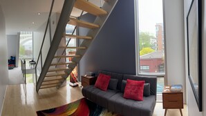 Uma sala de estar moderna com escada de madeira, sofá cinza com almofadas vermelhas, uma janela grande e vista para o exterior.