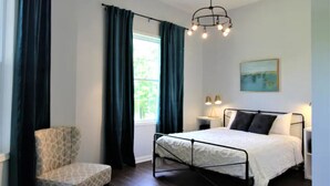Ein Schlafzimmer mit einem Bett, einem Sessel, einem Nachttisch mit Lampen, einem Fenster mit Vorhängen und einem Gemälde an der Wand.