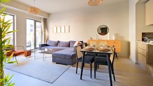 Een moderne woonkamer met een bank, een eettafel met stoelen, een keuken en grote ramen.