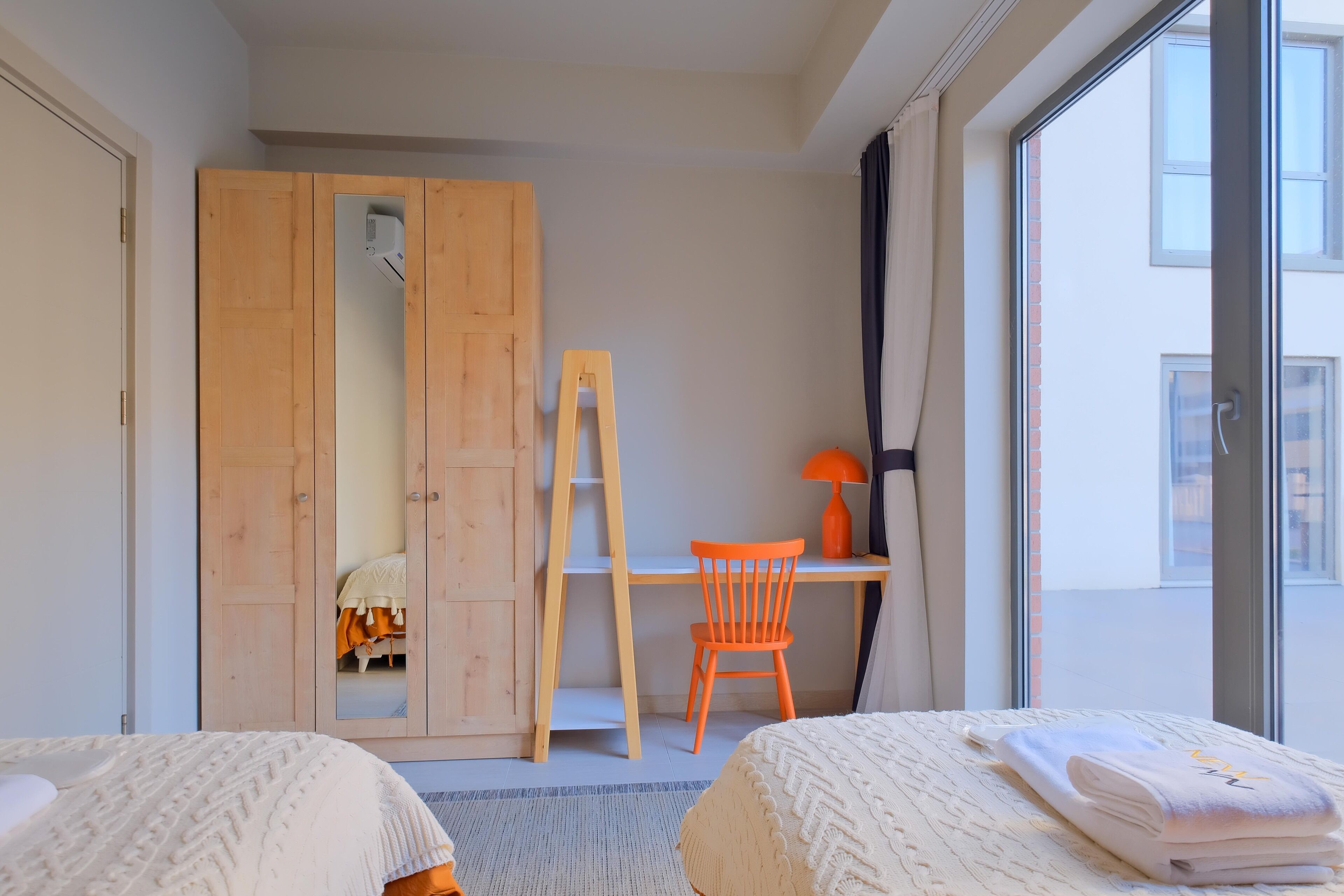 Une chambre avec deux lits, une armoire en bois, un bureau avec une chaise et une fenêtre donnant sur des immeubles.