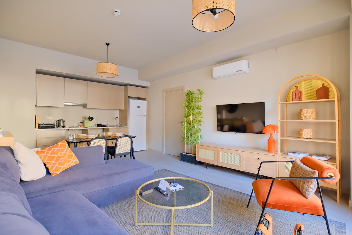 A modern living room with a sofa, coffee table, and orange armchair. A television and bookshelf are visible in the background.