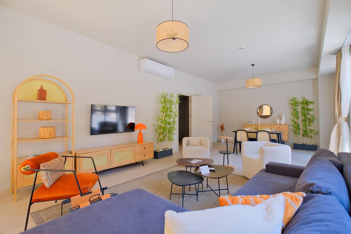 A modern living room with a sofa, armchairs, a coffee table, and a TV. There are plants, a dining area, and a kitchen in the background.