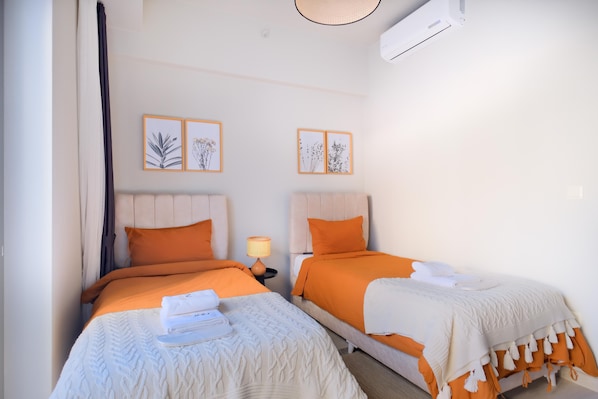 A hotel room with two beds, each with orange bedding and white headboards. There are framed botanical prints on the wall, a bedside lamp, and a white air conditioning unit. - NEW INN Hotel Cadde 54 Mall (Serdivan)
