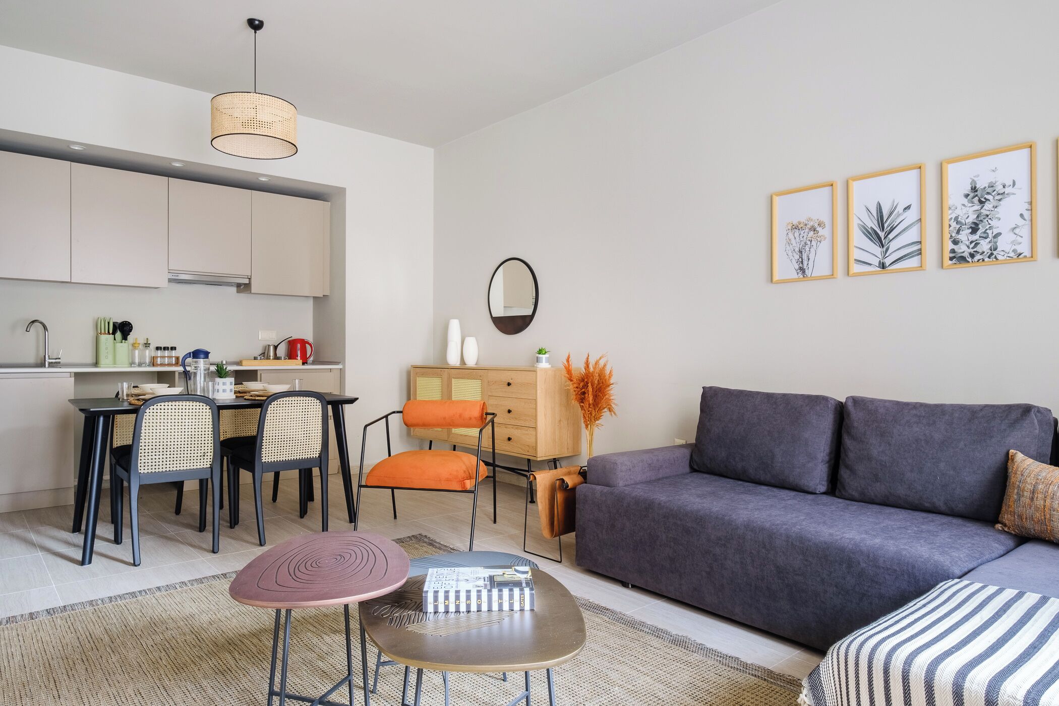 Un saló modern amb un sofà gris, una taula de dinar amb caires, una taula baixa i una zona de cuina amb armaris i un llum penjant.