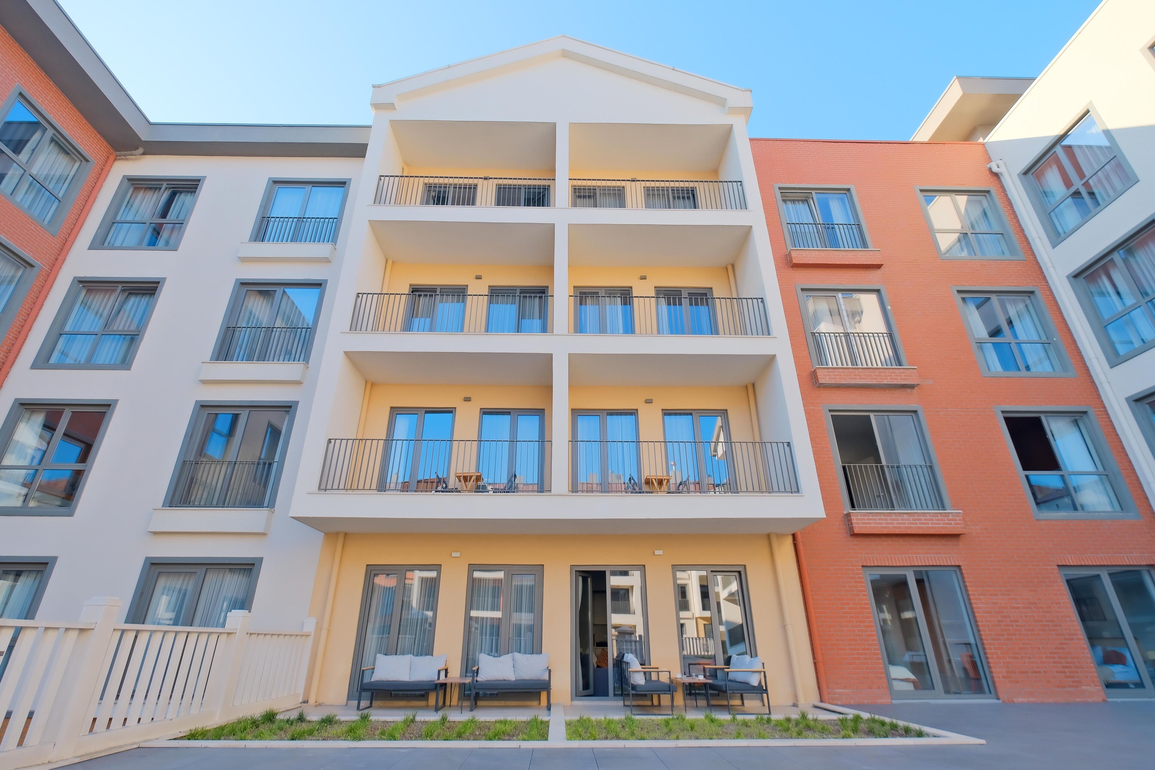 Un immeuble résidentiel à plusieurs étages, doté de balcons, d’une entrée sécurisée et d’un espace extérieur aménagé pour s’asseoir.
