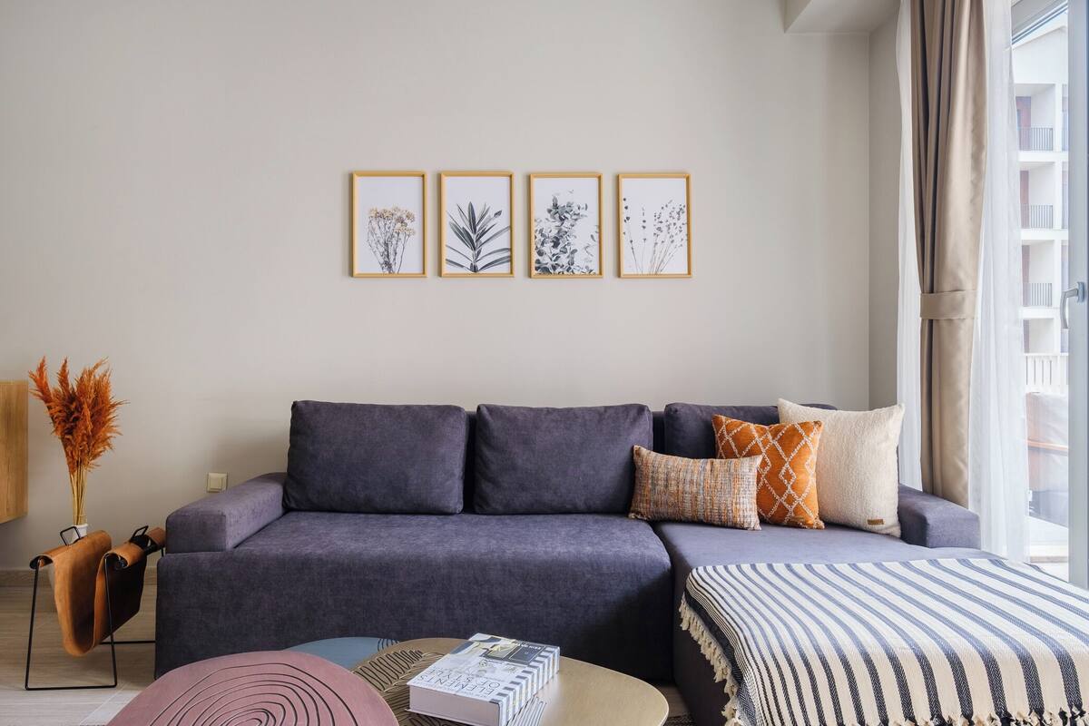 A modern living room with a grey sofa, a striped area rug, and a coffee table with books. There are framed botanical prints on the wall and a vase with dried flowers on a side table.