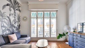 Un salon avec un canapé gris, une table basse, un meuble bleu et une grande fenêtre dotée d’auvents à rayures.