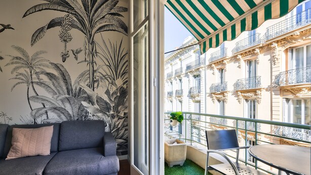 Un salon avec un canapé gris, un balcon recouvert de gazon synthétique vert et une vue sur un immeuble doté de balcons.