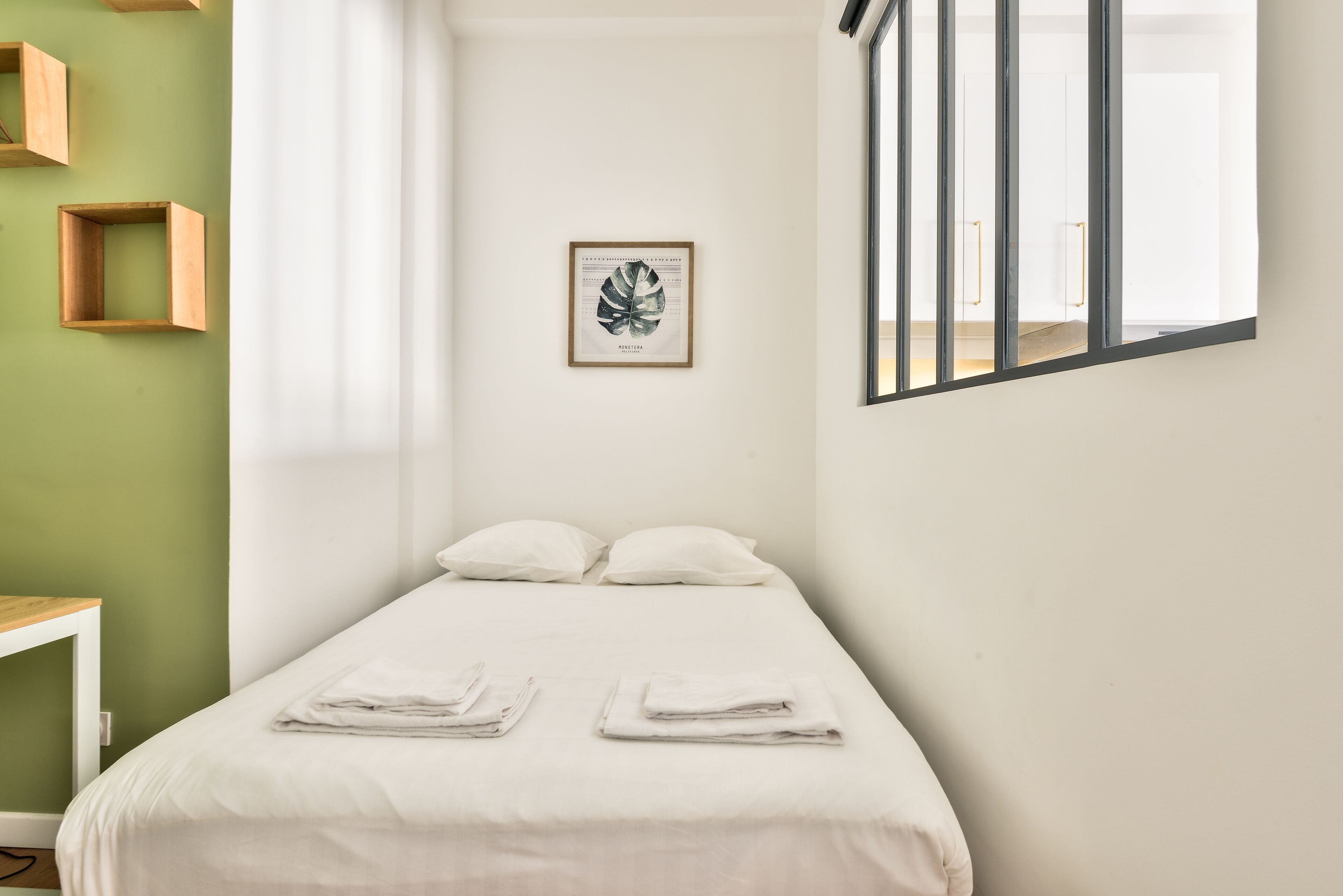A neatly made bed with folded towels, a framed botanical print on the wall, and a window with vertical blinds.