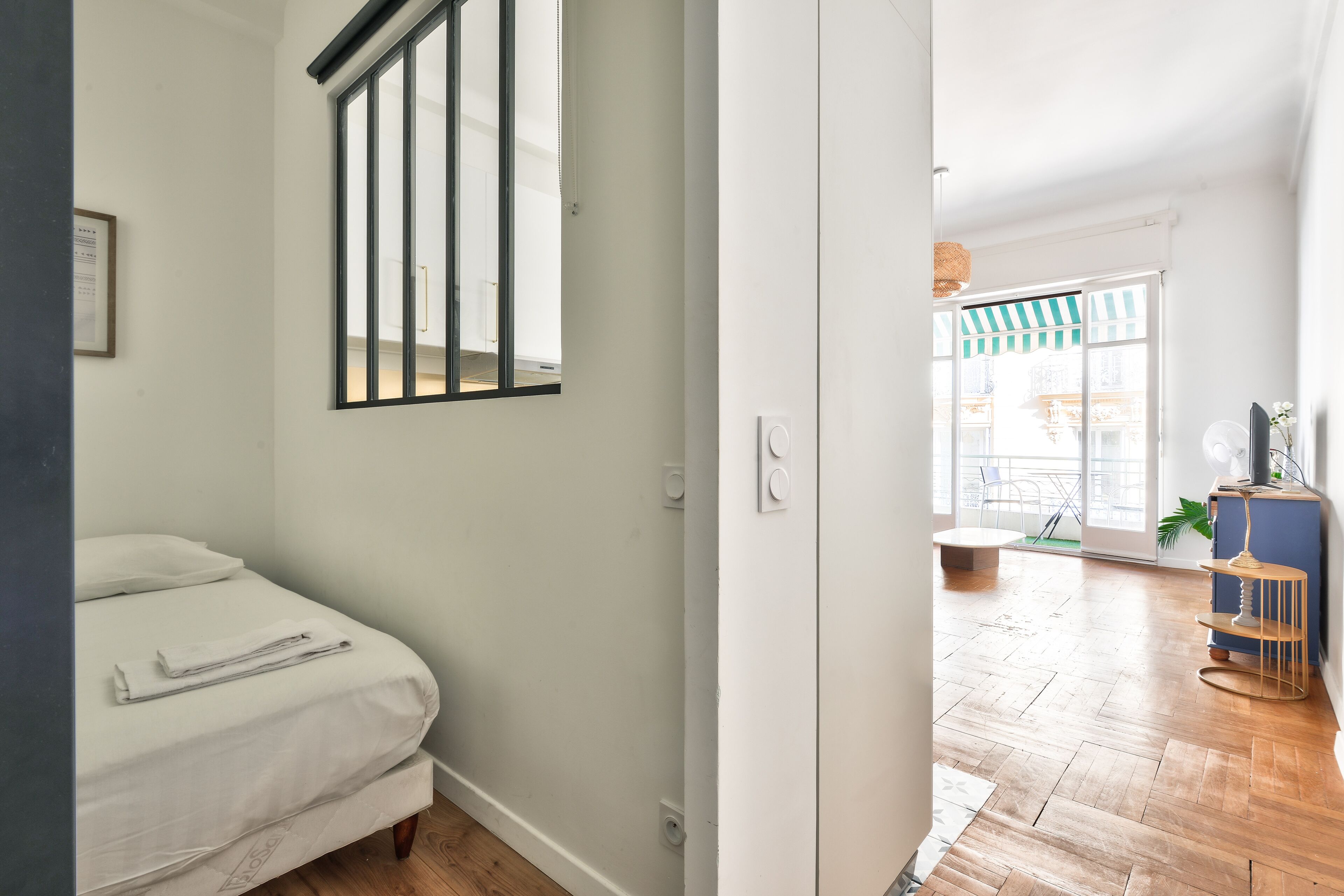 Een slaapkamer met een bed, een raam met jaloezieën, een deur naar een andere kamer en uitzicht op een buitenruimte met een stoel en een plant.