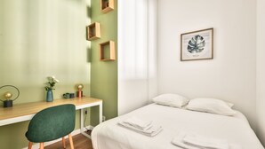 Une chambre bien rangée, avec un lit, un bureau et une chaise. On y trouve trois étagères en bois fixées au mur, une estampe botanique encadrée et un vase avec des fleurs posé sur le bureau.